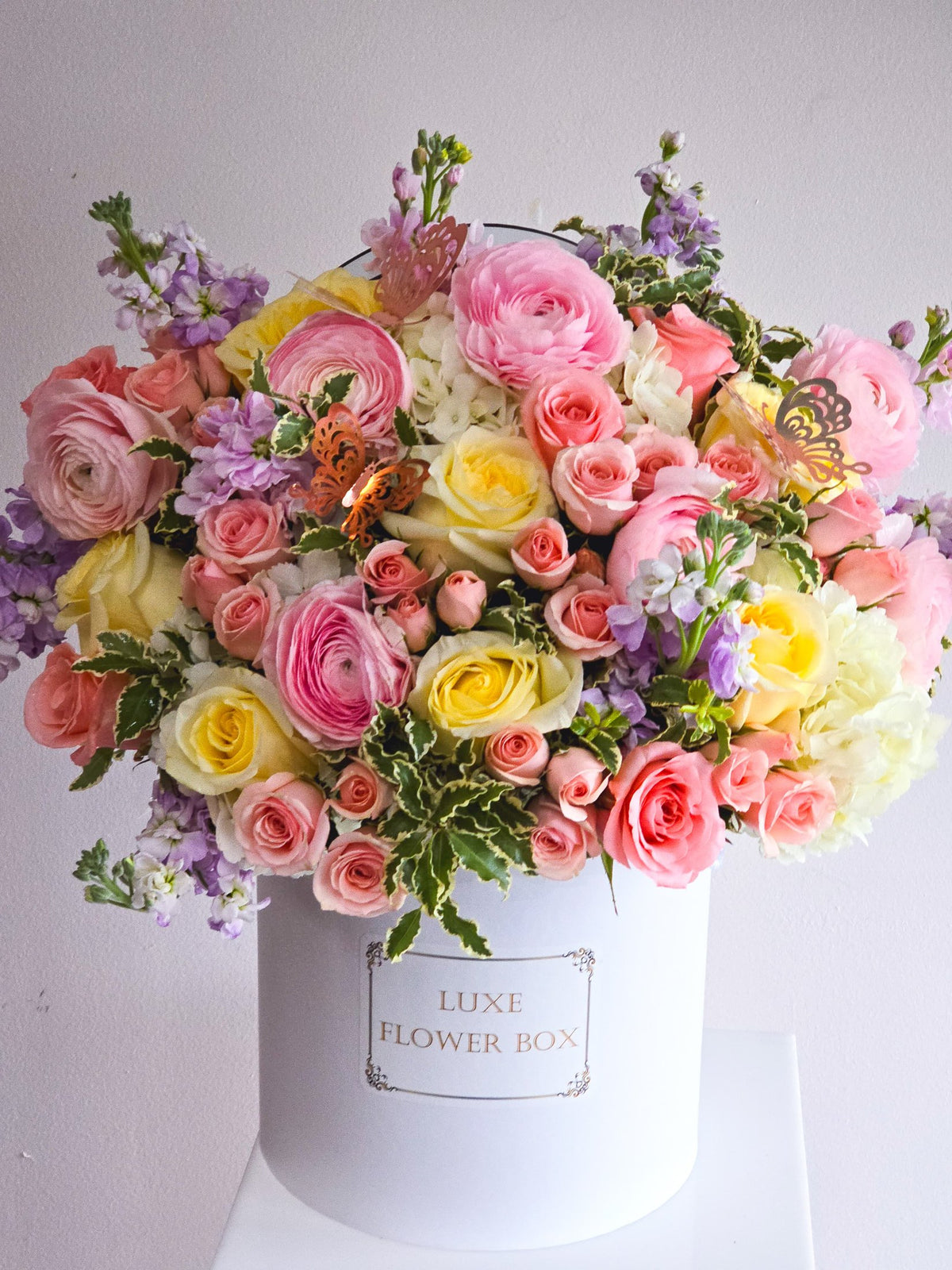 Chic flower box arrangement 'MIA' featuring lush greenery and vibrant blooms in yellow, peach, lavender, and white hues, including ranunculus, spray roses, roses, and stock for a fresh and elegant display.