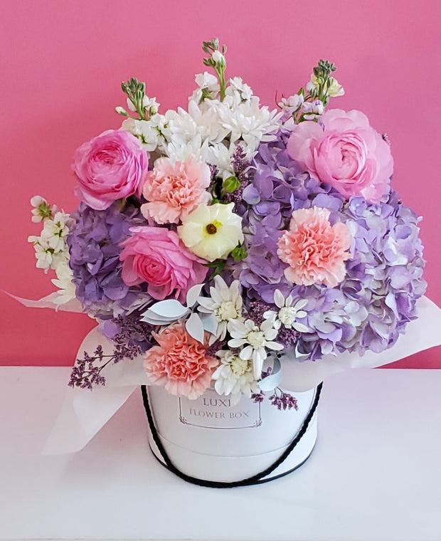 A floral arrangement featuring lavender hydrangeas, pink carnations, and ranunculus in a white box.